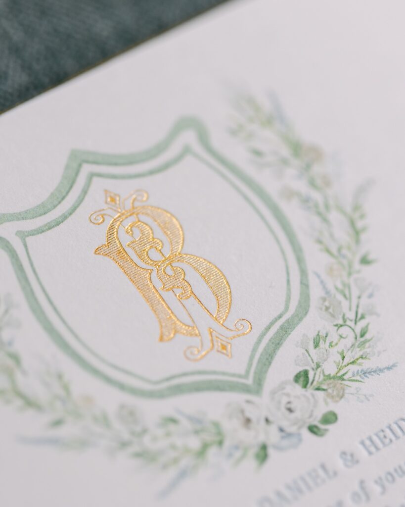 Gold foil monogram shield crest with watercolor flower border. Custom wedding invitation design. Laura Hooper Design House.